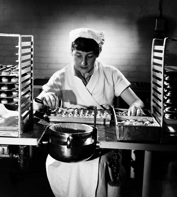 A black and white photo of a woman in a white uniform and cap at a workbench. She is dipping sweets into a squat metal pot with one hand while her other hand reaches for an almond to lay on top. There is a batch of assembled sweets on the bench, and two metal boxes containing undipped sweets and almonds. A patch of light behind her head creates a halo effect. She is flanked on either side by trays of completed sweets on tall racks.