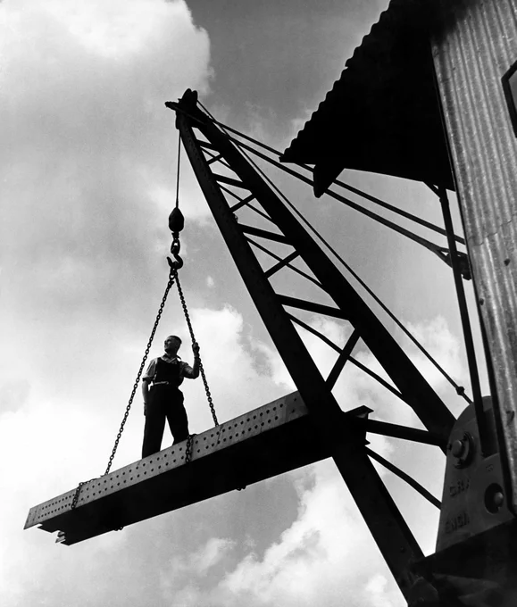 A black and white photograph of a man standing on a steel beam as it’s lowered from a tall crane. He is almost silhouetted against the sky, and stands casually with one hand in his pocket and the other holding the chain, looking up and out of the photo.