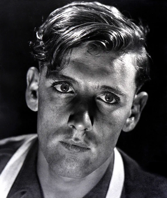 A high contrast black-and-white portrait photograph of a young man. His eyes are focused on a point off camera, just past the photographer. Light reflects off his hair, pushed back off his forehead, and the background is a deep black. The effect is closer to a portrait of a film star than a factory worker.