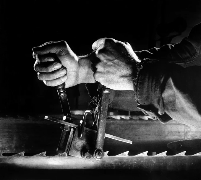 A black and white photograph of a pair of hands gripping a metal tool to adjust the teeth of a saw. The background is a deep black and the light highlights the shape of the saw teeth along the bottom of the frame.