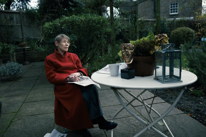 A image of actress, Glenda Jackson in a red coat. She is sat on a chair in a garden and there is a newspaper on her lap. She is sat by a garden table which has a BAFTA award on it as well as a mug, plant and lantern.
