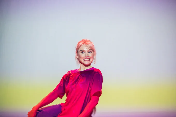 PRODUCTION SHOT: Alice sits smiling with her gaze to the left in a pink t shirt lit with a yellow warmth and a white backdrop 