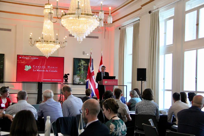 Greg MacDonald stood behind the podium on stage addressing a room full of people with the Canada House logo visible in the background.