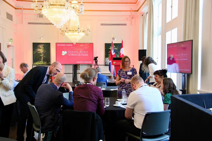 A group of people in conversation around a table with the Canada House logo visible in the background.