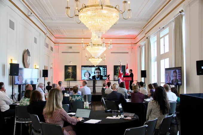 Room full of people looking at screens where virtual participants are joining the conversation, with Canadian and UK flags in the background.