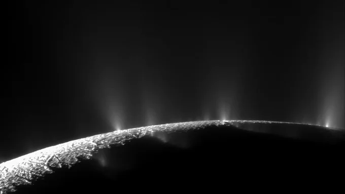 In shades of black and white, thin plumes spray upwards from the surface of a bumpy moon. The moon is seen from space, curving slightly downwards from right-to-left across the image; its foreground is mostly in shadow. Credits: NASA/JPL-Caltech/Space Science Institute