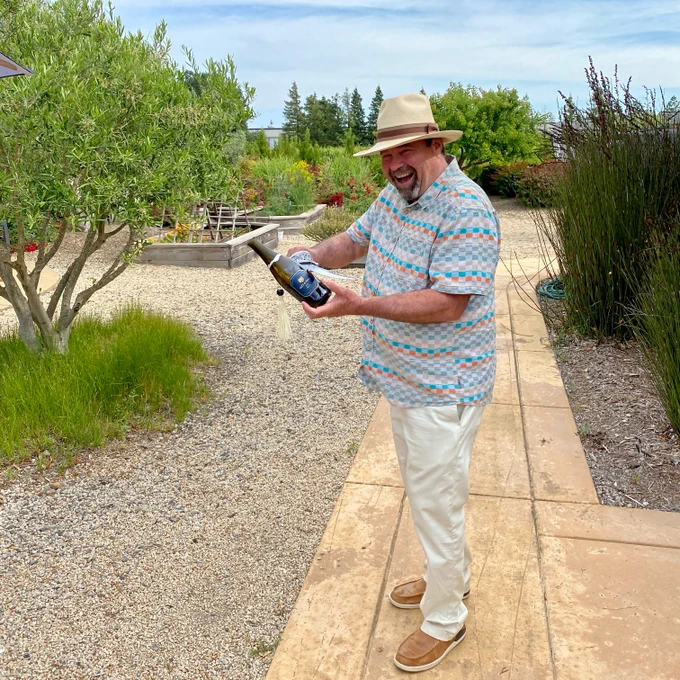 Patz & Hall Winemaker James Hall sabering a bottle of sparkling wine outside at the Sonoma House tasting room. He is wearing light summer clothes and a hat. 