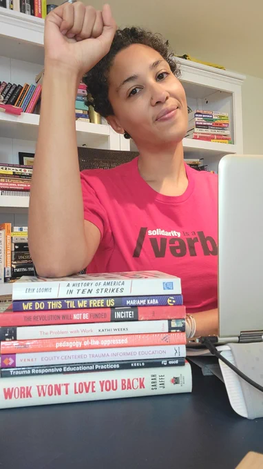 Cee in front of open laptop. My arm rests on stack of books about labor, justice and trauma and leads up to raised fist. My shirt is red - my staff union's color - and reads "Solidarity is a Verb." I have a light smile on tilted head. Behind me are full bookshelves.