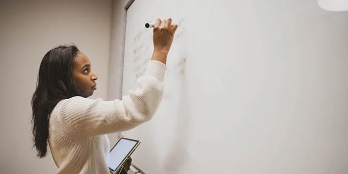A woman in her early 20s stands in front of a dry erase board writing on it, she's holding an Ipad in her other hand.