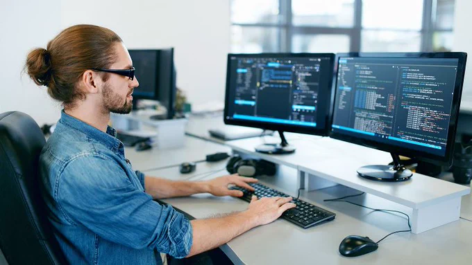 Man with bun working at desk on computer with two screens