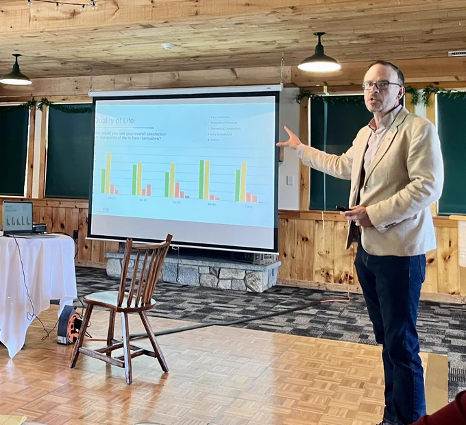 image shows a man speaking, with a presentation screen in the background