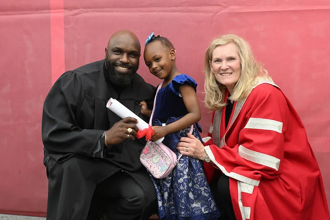 York President Rhonda Lenton poses crouching down with a School of Continuing Studies Graduate and their daughter in front of a red picture wall following the SCS graduation ceremony.
