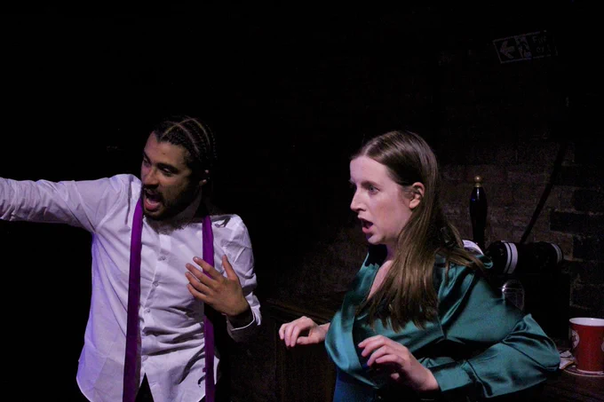 Isah, a young mixed race Black man, sat with a loose tie and a white shirt, stood at a bar, next to Brontë, a young white woman with long light brown hair and a green dress. They are both looking shocked to the left of frame.