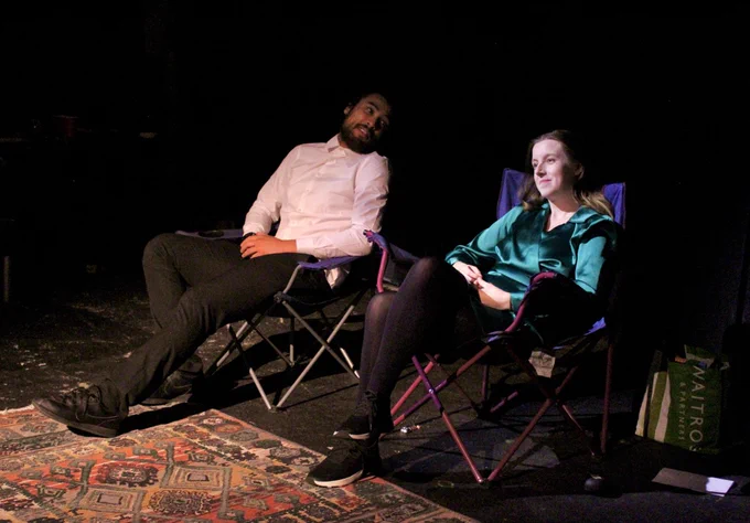 Isah, a young mixed race Black man, sat with a white shirt, with Brontë, a young white woman with long light brown hair and a green dress. They are both sat on camp chairs and laughing gently together,