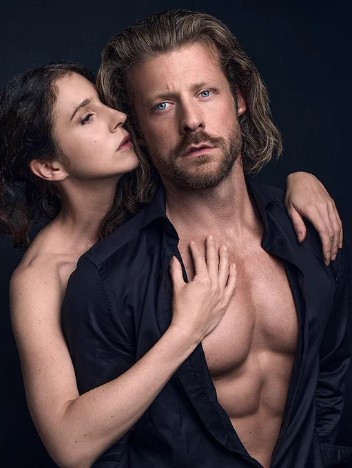 Jessica Boone (Juliet) standing behind and with her body pressed against Taylor Napier (Romeo). Jessica's bare arms are wrapped around Taylor, and she is touching his bare chest with her right hand. Taylor looking at camera with a black long sleeve shirt opened wide and unbuttoned to the waist.