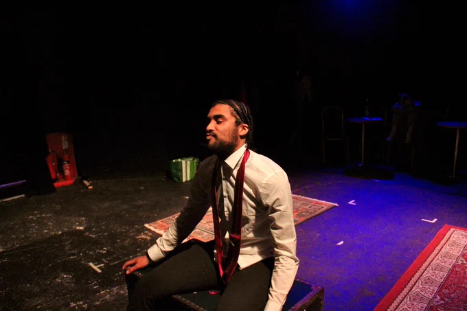 Isah, a young mixed race Black man, sat with a loose tie and a white shirt, on a blue trunk in the middle of a stage. Behind him are two carpets.