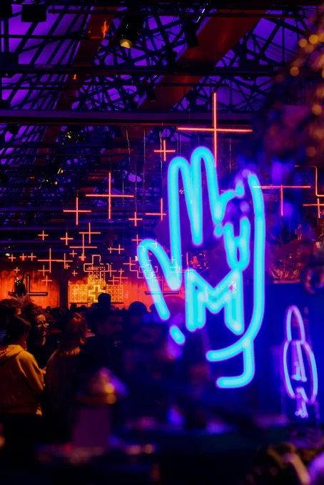A bright blue neon light of a hand gesture with Moorilla Winery's "M" and a smaller neon sign of a club (like on playing cards) with Domaine A Winery's "A" are out of focus in the foreground of the photo, with the red neon crosses of Dark Mofo's Winter Feast in focus behind them.