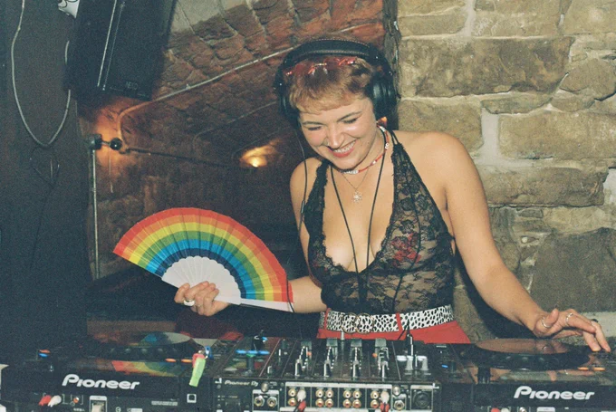 A film photograph of Femmergy DJ Annafleur holding a rainbow fan behind a DJ deck