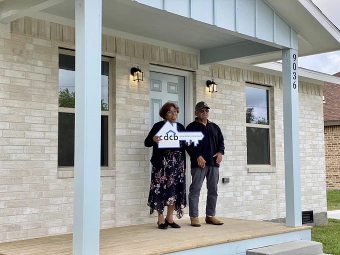 Gloria and Evelio Palacios standing proudly in front of their new home. Gloria is holding a sign shaped like a key that says "cdcb". 