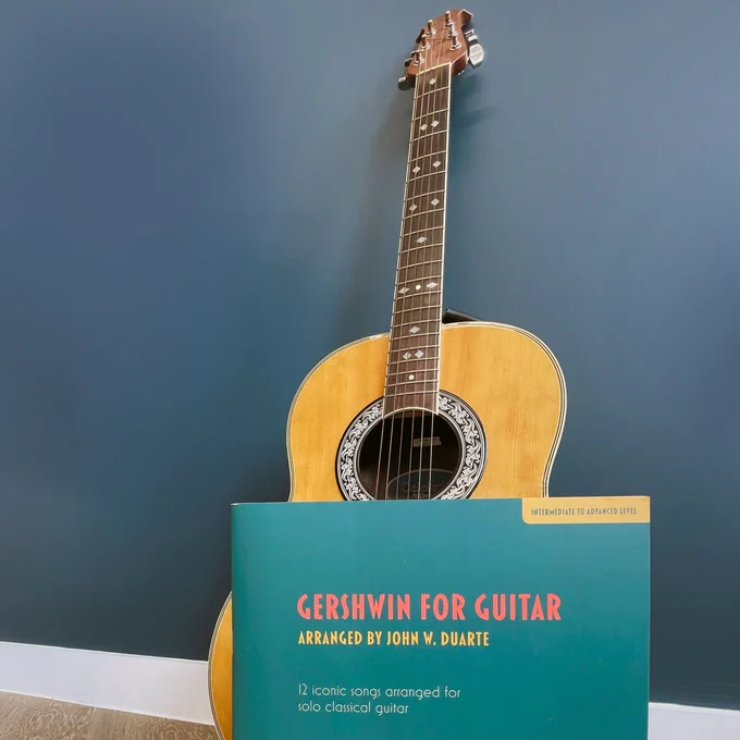A guitar leaning against a dark blue wall with the book 'Gershwin for Guitar' in front