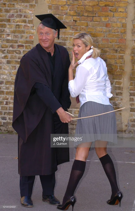 Chris Tarrant is seen dressed as a schoolmaster, he is holding a cane and hitting Nell McAndrew (dressed in a grey skirt, white blouse, and long socks) on the backside with it. They are stood in a school playground.