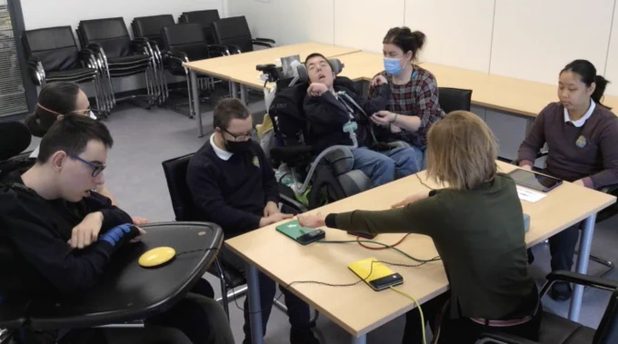 Photo shows from left to right around a table in a classroom - a boy wearing navy school uniform in a wheelchair playing a yellow switch and his support assistant next to him: a boy in a mask and glasses in school uniform playing a green switch; a boy in a wheelchair in school uniform playing a blue switch with his elbow held by a female teacher in a tartan top wearing a mask; and a female student in school uniform playing iPad. All of them have dark hair. In front of the image is DMS musician Abi wearing a green top with fair hair helping one boy with his switch. 