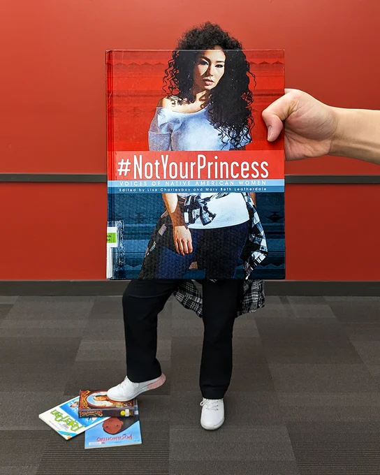 Evoking the cover of the book #NotYourPrincess: Voices of Native American Women, edited by Mary Beth Leatherdale and Lisa Charleyboy, a young woman stands in front of a red wall with her right foot on top of books about Peter Pan and Pocahontas.