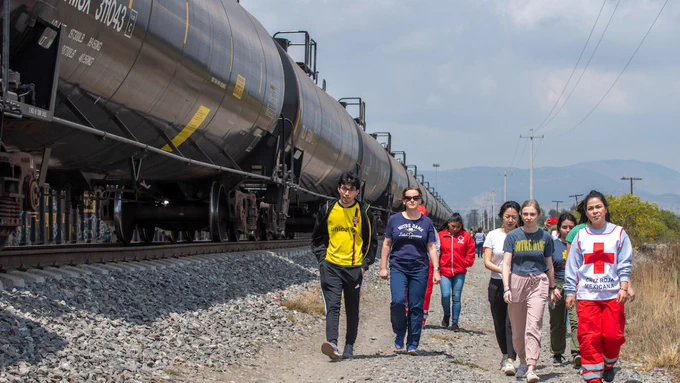 Men and women follow a woman wearing a Red Cross vest and red pants as they walk alongside a freight train in Mexico.