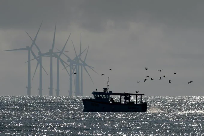 Overcast day with mostly gray and silver tones. In the background at left, a line of 8 offshore windmills tower over the horizon. In the foreground, a small open-decked boat with a flat roof is silhouetted against a shining sea.