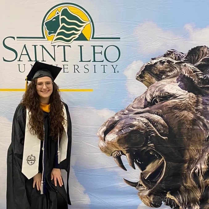 Lace smiling in graduation regalia in front of a banner labeled ‘Saint Leo University’
