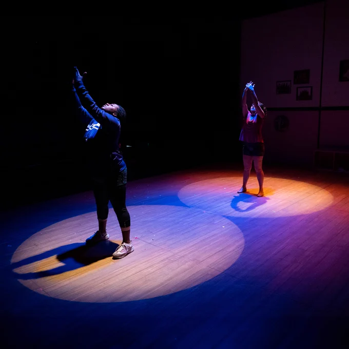 A photo of actors Kylah D'Jai and Natalie Hanson, each standing in a circular pool of light, their hands outstretched above their heads in a dance pose. Dark blue and purple lights illuminate the edges of the space, creating a cool, mellow atmosphere.