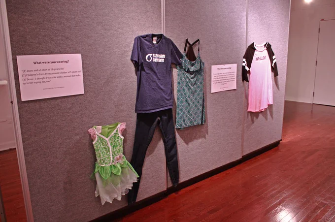 a variety of clothes in different sizes including a t-shirt and jeans and a child's tinkerbell dress hung up on the wall. there are signs that say "what were you wearing" and describe what each person was wearing when they were sexually assaulted. the first sign lists the following:

1. jeans and a t-shirt at 18 years old
2. children's dress, by my father at 5 years old
3. dress - i thought i was safe with a woman, but woke up to her raping me too