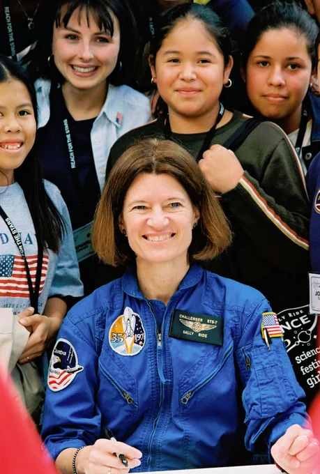 Dr. Sally Ride in blue flight suit, seated, at NASA outreach event with four young women with lanyards standing behind her as they smile for a photo.
