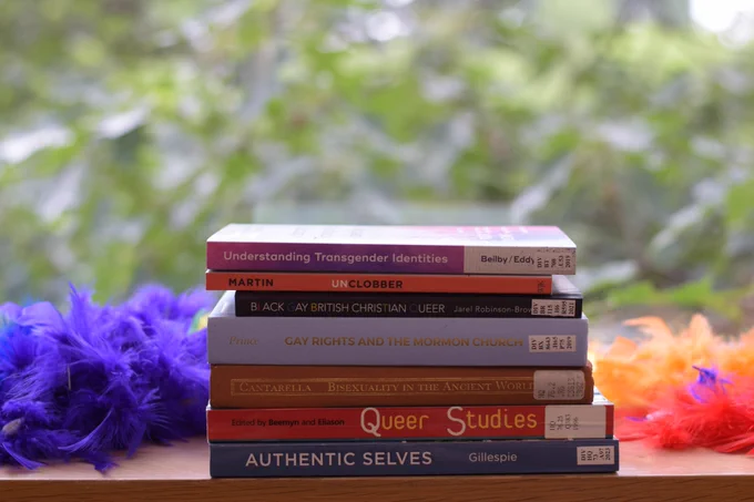 Several books related to LGBTQIA  month sit on a windowsill. A rainbow boa is draped behind the books.