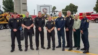 Eight people in uniform are arranged in a semi-circle and looking at the camera. Large equipment, including a red fire engine, is staged behind them. 