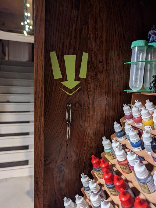A very bright green arrow points down to two paperclips linked together next to some tiny paint bottles.