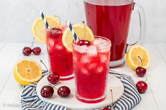 Two clear glasses filled with a bright red lemonade with lemon slices on  the side.