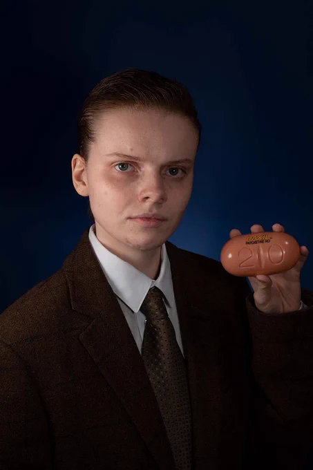 A photo of a person dressed in a suit and tie, holding a pill box and looking straight into the camera. Their hair is slicked back and the image feels cooperate. Artwork by Ruby Cromer.