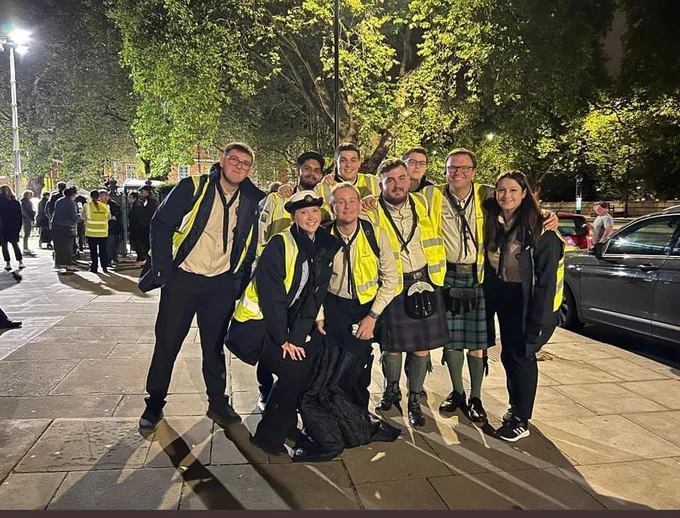 A group of smiling Scouts volunteers in hi visibility jackets, lit by a streetlight, in front of the queue for the Lying-in-State of Queen Elizabeth II.