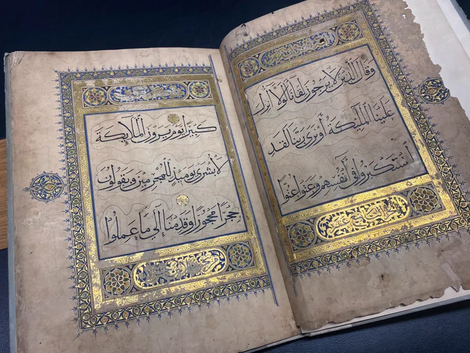MS Mocatta 20 open to show two pages at the beginning of the manuscript. It features clear Arabic script at the centre of each page, surrounded with an intracate blue and gold leaf border. The light shines off of the gold on the bottom of the page. 