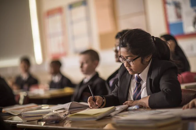 School girl sitting in class writing at a desk, other pupils can be seen in the background out of focus.