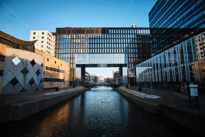 Picture of the UvA building at Roeterseilandcampus. It is showing the building from the water. You have a view of the canal going under the black, shiny building. On the left of the canal is a beige building and on the right there is another shiny black building. The sky is a clear blue color. 