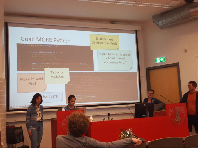 A group of people in front of a presentation in a lecture hall. Slide title: Goal: MORE Python