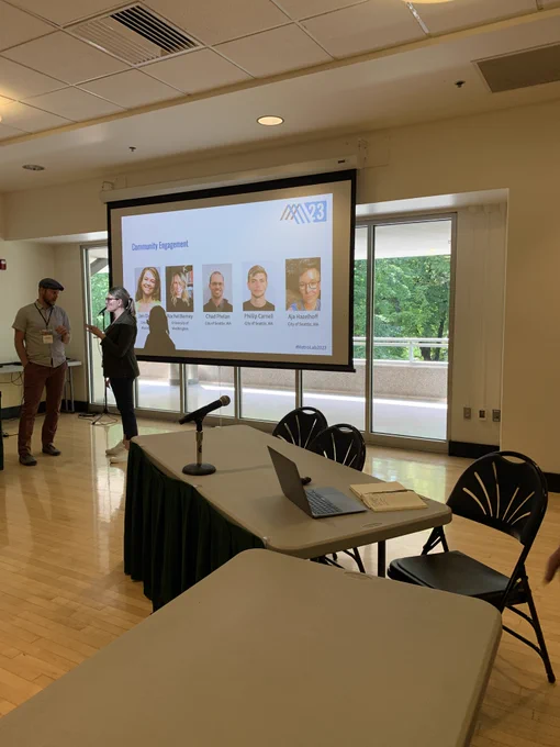 A photo of a screen that has a projected image of the presenters for the community engagement panel, which includes those from the City of Seattle (Chad Phelan, Phillip Carnell, Aja Hazelhoff).