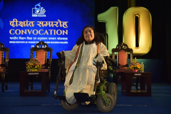 Shireen, in a white dress sitting in her wheelchair, smiling gently at the camera. A white shawl with a red and green border is on her shoulder. The background is blue, and the text says CONVOCATION, IISER Kolkata.