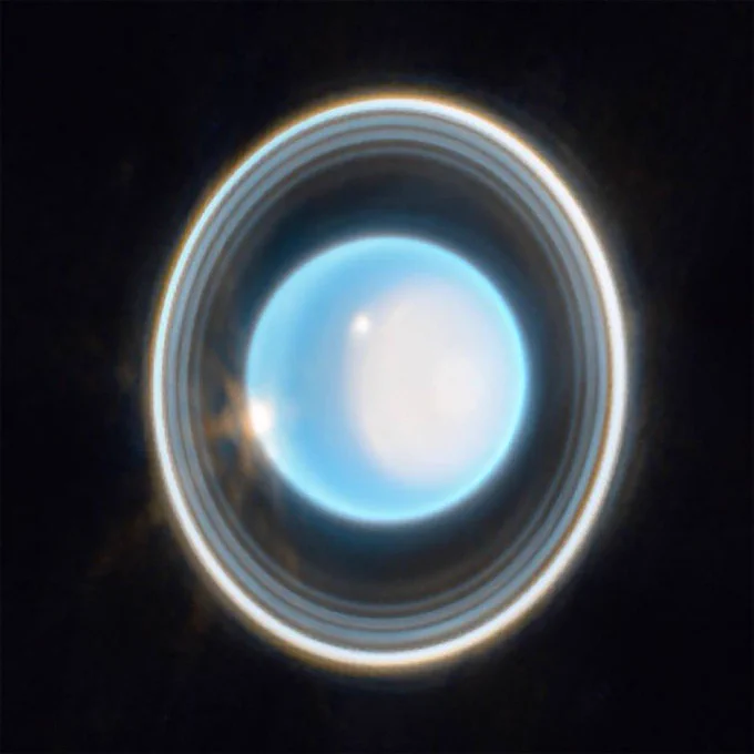 The planet Uranus on a black background. The planet appears light blue with a large, white patch on the right side. On the edge of that patch at the upper left is a bright white spot. Another white spot is located on the left side of the planet at the 9 o’clock position. Around the planet is a system of nested rings. The outermost ring is the brightest while the innermost ring is the faintest. Unlike Saturn’s horizontal rings, the rings of Uranus are vertical and so they appear to surround the planet.