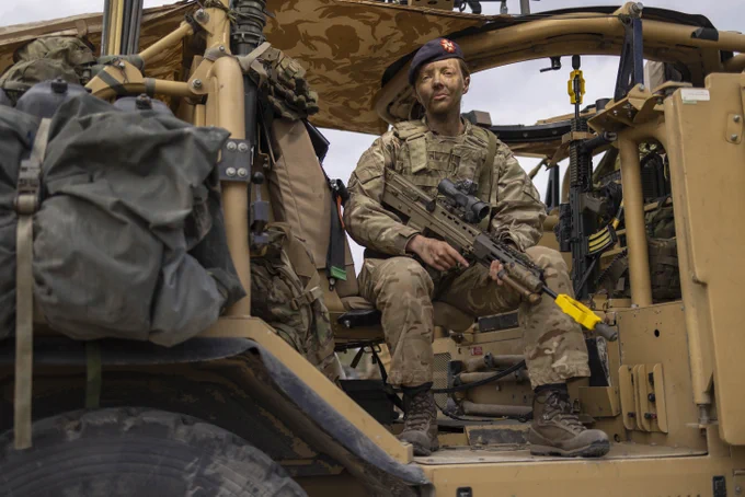 A female British Army officer is sitting inside a military vehicle. She is wearing camouflage uniform, a dark blue beret on her head, and holding a rifle right to left, across her chest. 