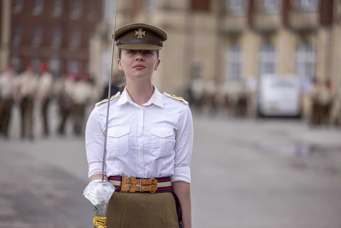 A female British Army officer is standing on a parade square facing the camera. She is holding a sword upright in her right hand. Also, she is wearing a short sleeved shirt, a khaki coloured skirts and hat. 
Blurred out in the background are soldiers standing in front of a large building. 