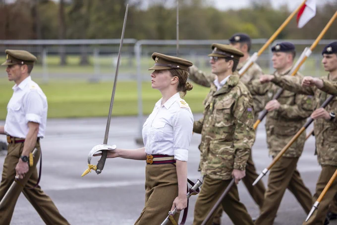 British Army male and female soldiers are marching in rows along a road.  The two officers, one male and one female are holding swords upright in their right hands. The soldiers behind are carrying cavalry lances in their left hands sloped against their shoulders. The officers are wearing short sleeved shirts, and khaki coloured skirts and trousers, the soldiers are wearing camouflage jackets.