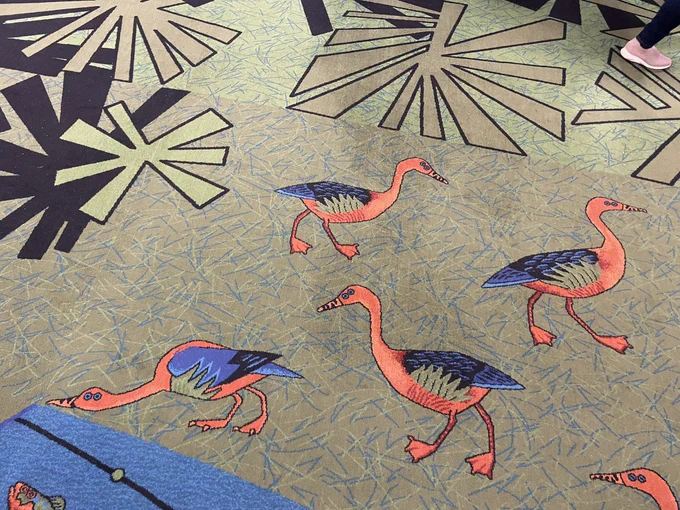 Photo of a carpeted floor with a pattern of four ducks on it. They have orange bodies, blue wings, and pale blue plumed pants.